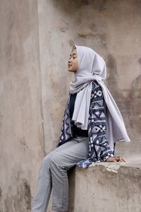 Young woman with eyes closed wearing hijab sitting against wall