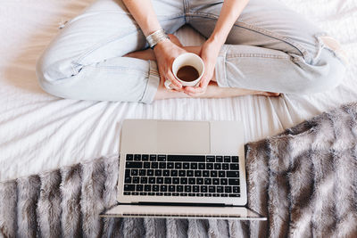 Low section of woman holding coffee while sitting by laptop on bed