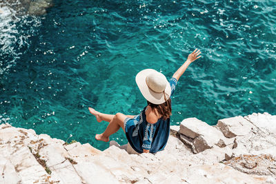 Young woman sitting on a cliff above sea, arms raised, happy, adventure, summer, vacation.