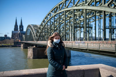 Portrait of young woman wearing mask standing against bridge over river