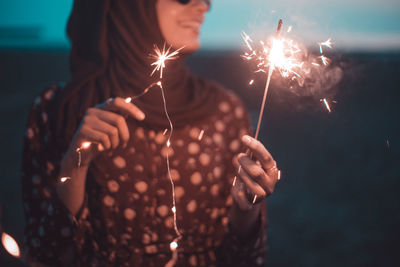 Young woman with illuminated sparklers standing at beach during dusk