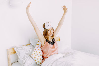 A happy cute girl in pink pajamas and a sleep mask wakes up and stretches on the bed at home