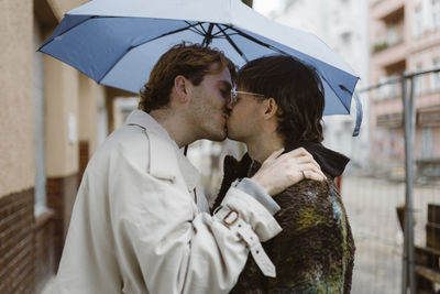 Side view of affectionate gay couple kissing under umbrella at street