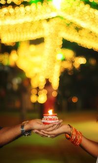 Cropped hands of couple holding lit diya against illuminated lights at night