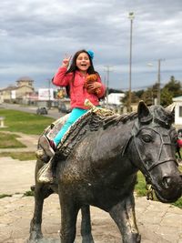 Happy girl showing peace sign while sitting on horse sculpture