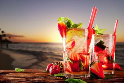 Fruits and drink in glasses on table at beach against sky during sunset