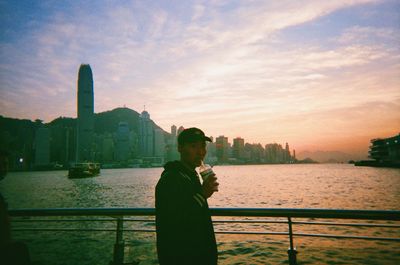 Man standing by railing against river and buildings against sky during sunset