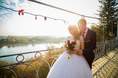 Bride and groom standing on bridge over lake and sky on sunny day