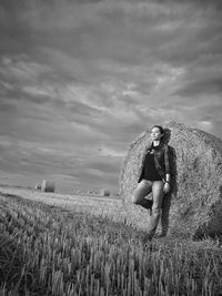 Young woman looking away while standing by hay bale against cloudy sky