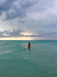 Rear view of young woman wearing bikini while standing in sea against cloudy sky