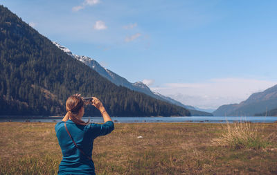 Rear view of woman photographing standing on grassy field against blue sky