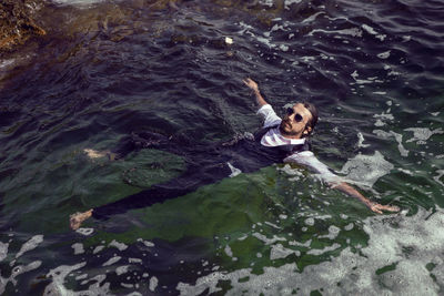 Man with a beard and sunglasses in clothes a vest and a white shirt swims in the sea among the rocks