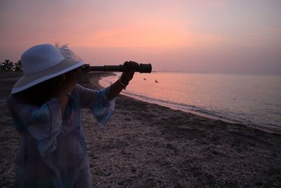 Woman looking through binoculars at beach against sky during sunset