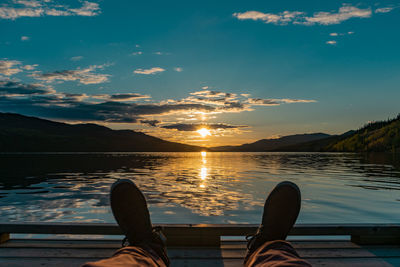 Low section of person by lake against sky during sunset