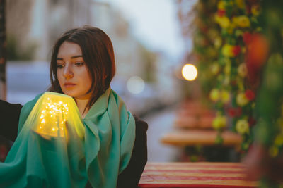 Thoughtful beautiful woman holding scarf and glowing jar in city at dusk