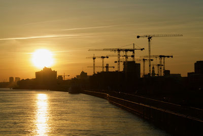 Silhouette buildings by river against sky during sunset