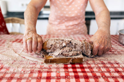 Anonymous old female rolling out dough on cutting board at table in house