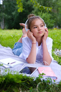 Teen girl lies listens to music in headphones on the grass in the park with books and notebooks