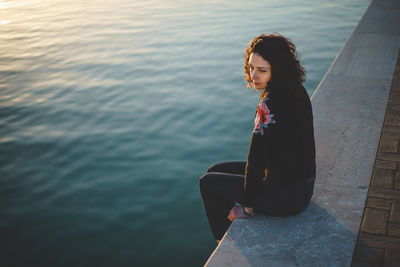 Woman sitting on retaining wall over sea during sunset