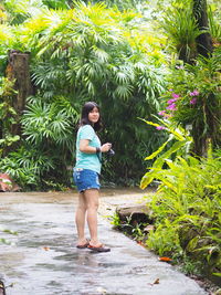 Portrait of woman holding camera standing on footpath against plants