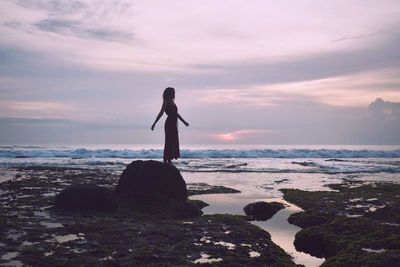 Woman standing on rock at beach against sky during sunset