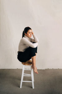 Side view of young unhappy barefoot female in casual clothes sitting on stool and looking away