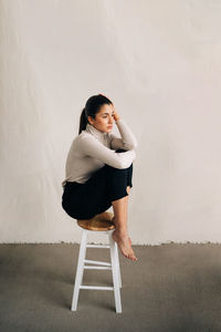 Side view of young woman sitting on chair against wall