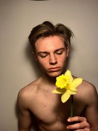 Shirtless young man holding yellow daffodil against wall