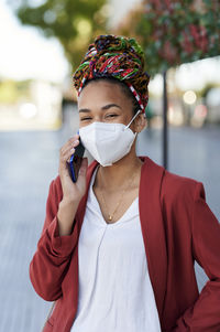 Young woman wearing headscarf and protective face mask talking on mobile phone while standing outdoors