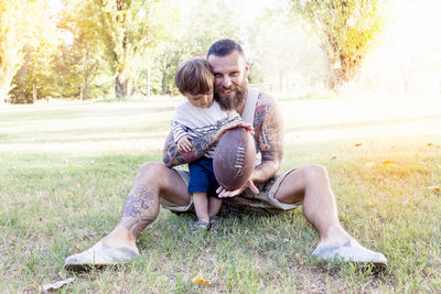 Portrait of smiling bearded man with son holding american football ball while sitting on grassy field