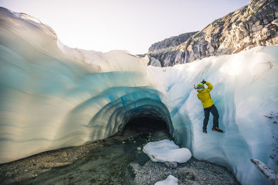 Man ice climbing next to glacial cave.