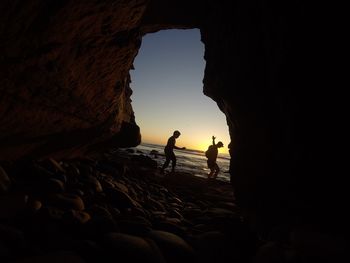 Silhouette couple standing by cave in sea against sky during sunset