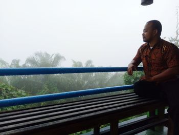 Man looking away while standing by railing against sky