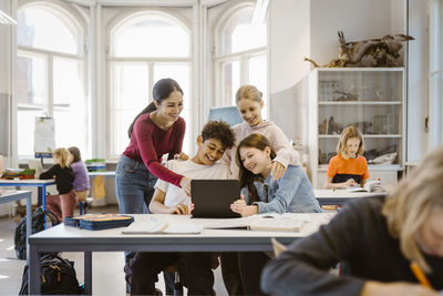 Smiling male and female students sharing tablet pc with teacher in classroom