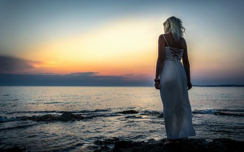 Rear view of woman standing on shore against orange sky