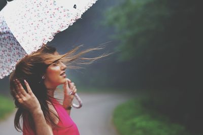 Side view of woman with tousled hair holding umbrella against trees