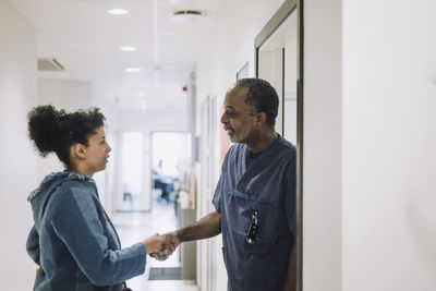 Smiling doctor greeting female patient with handshake while standing at doorway in hospital