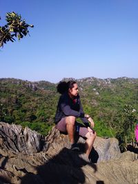 Young man sitting on rock against clear sky