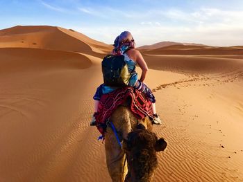 Rear view of woman riding camel at desert against sky
