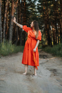 Barefoot happy young woman in red dress with hand raised dancing in pine forest at summer day.