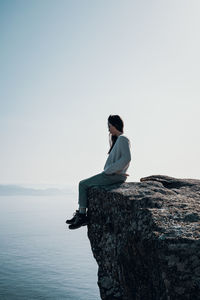 Woman sitting on rock against clear sky overlooking the ocean