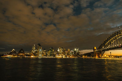 Illuminated bridge over river by buildings against sky at night
