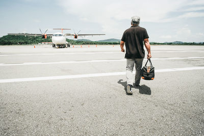 Rear view of man carrying bag while walking on airport runway against sky during sunny day