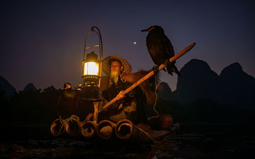 Man with lantern sitting by bird on boat in lake against sky during sunset