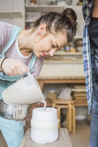 Young female potter pouring clay from pitcher in vase on stool by woman standing at workshop