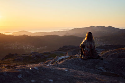 Rear view of woman sitting on rock against sky during sunset