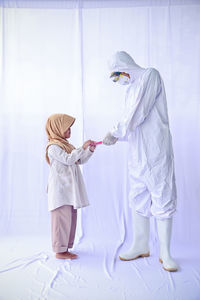 Full length of man wearing protective suit holding mask standing with cute girl against white background