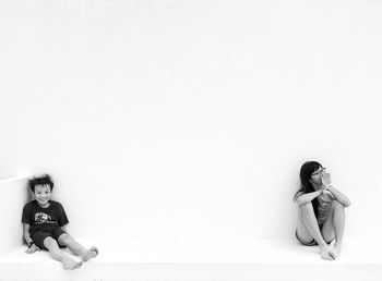 Full length of siblings sitting by white wall