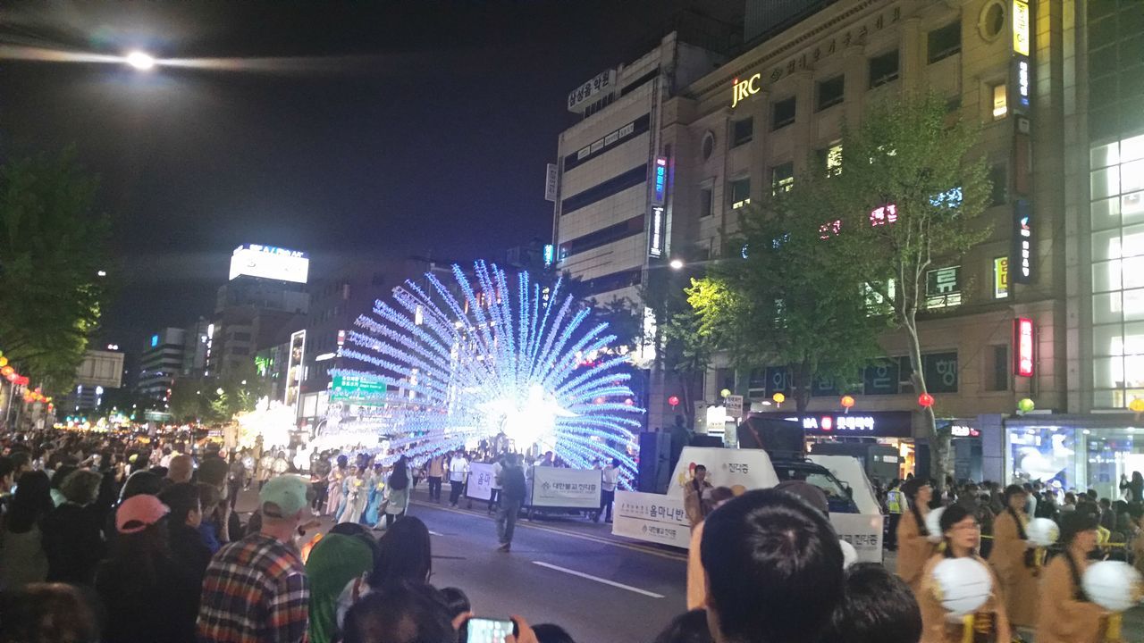 illuminated, large group of people, night, building exterior, city, architecture, built structure, crowd, city life, street, lighting equipment, men, city street, lifestyles, light - natural phenomenon, person, celebration, sky, leisure activity