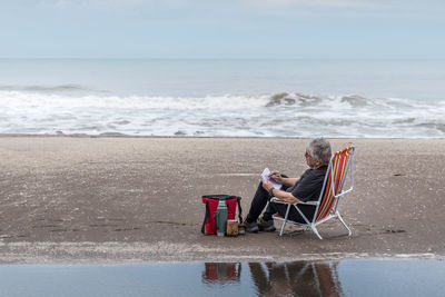 Adult man with gray hair sitting on a beach chair drawing. behind the beach and waves of the sea.